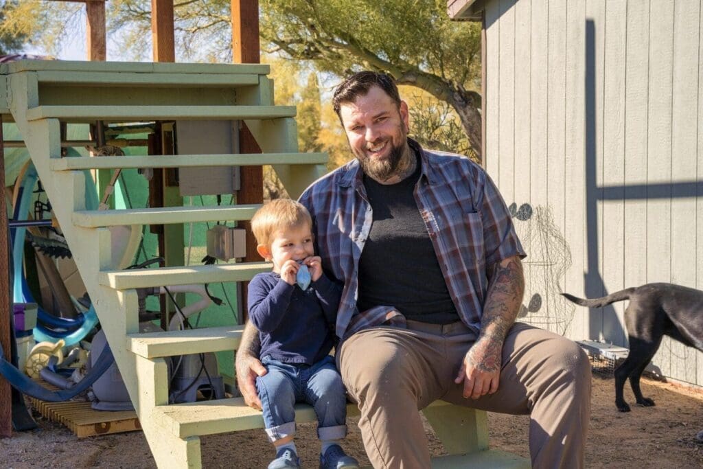 A man sits on painted outdoor steps with a young child beside him and dogs moving through the background.