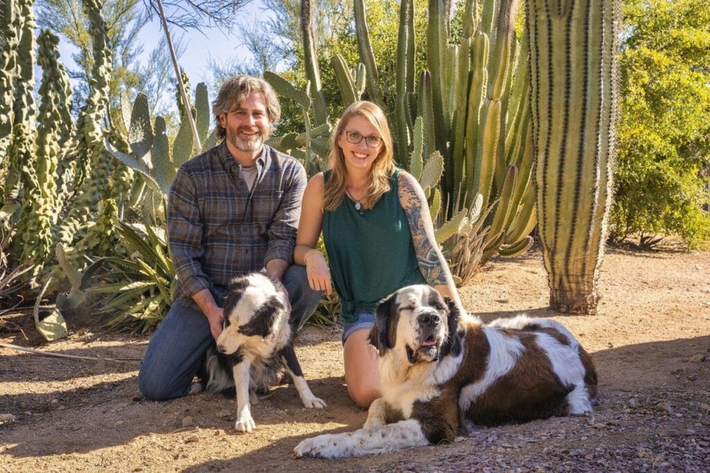 Two adults kneel beside two dogs in front of tall columnar cactus.