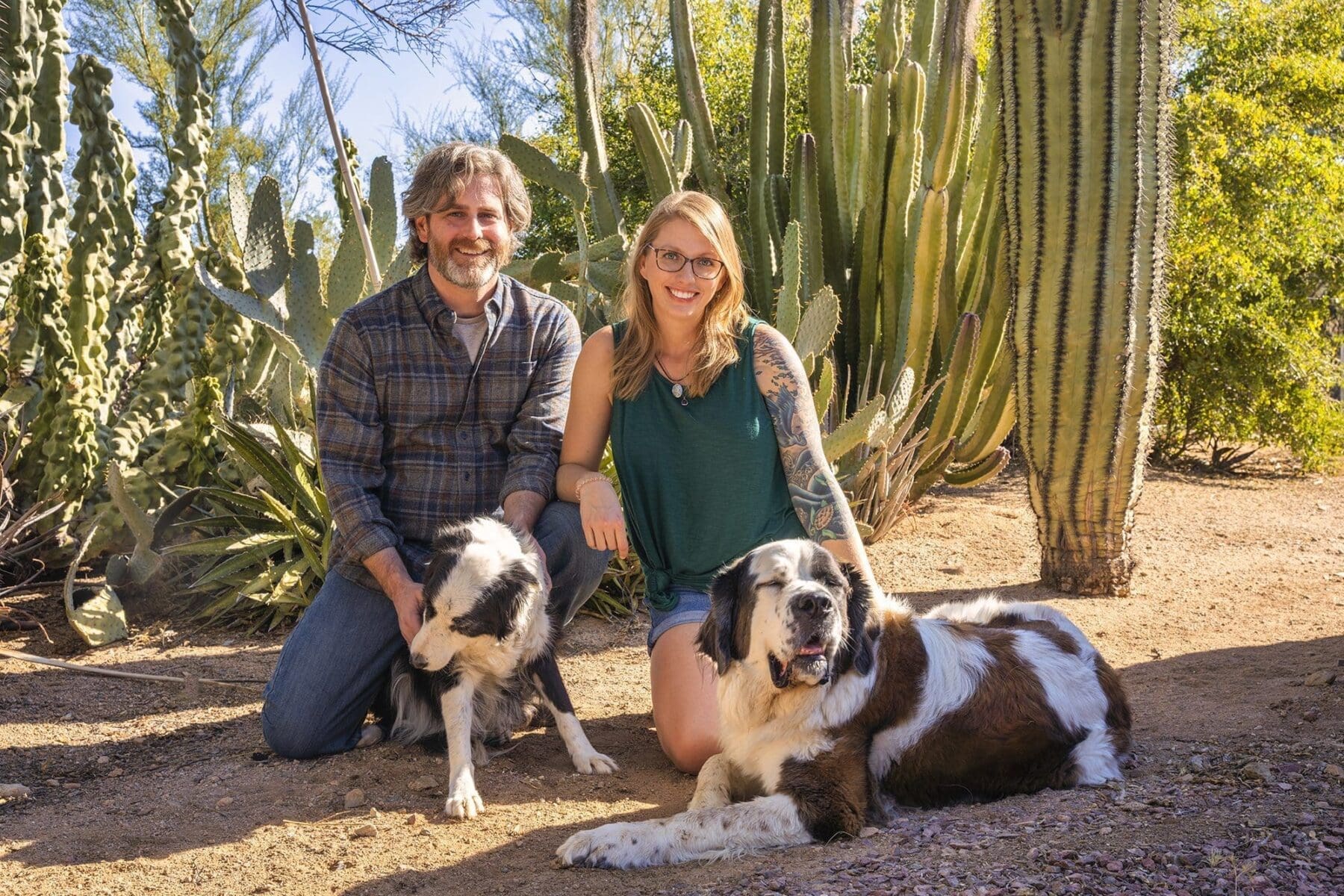 Two adults kneel beside two dogs in front of tall columnar cactus.