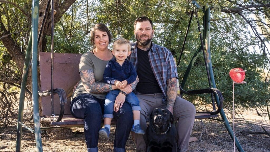 Two adults and a young child sit together on an outdoor swing with a black dog nearby.