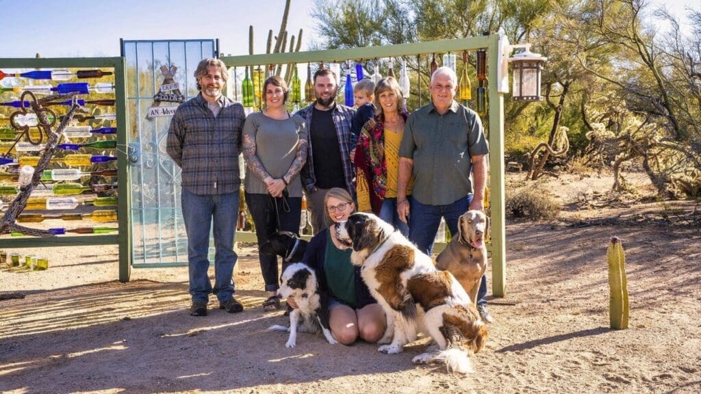 A large group of adults and a young child pose with several dogs in front of a colorful outdoor wall installation.