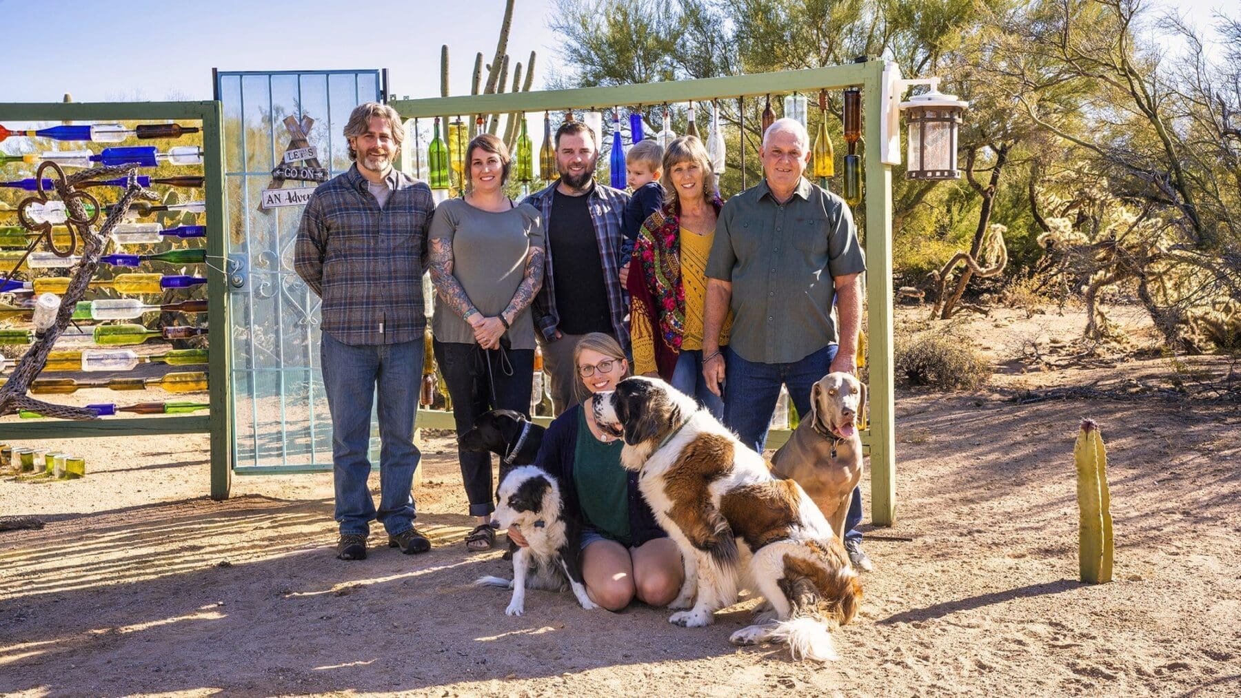 A large group of adults and a young child pose with several dogs in front of a colorful outdoor wall installation.
