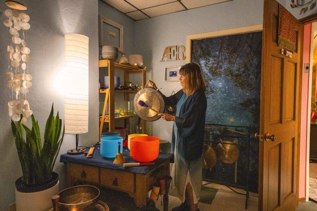 Kat Styrcula plays a metal gong indoors among sound-therapy bowls and instruments.