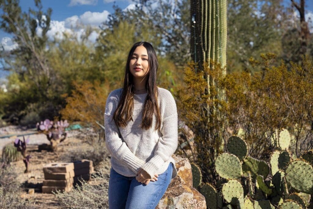 A young woman from the Simpkins family stands in a gray sweater beside prickly pear and a tall saguaro.