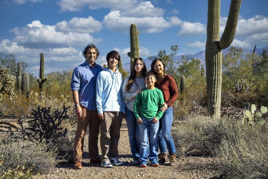 Five members of the Simpkins family stand together between tall saguaros beneath a cloud-filled sky.
