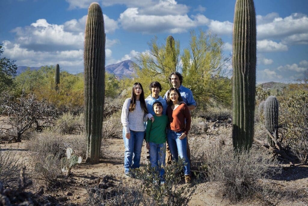 The Simpkins family stands together between two tall saguaros with a mountain and blue sky beyond.