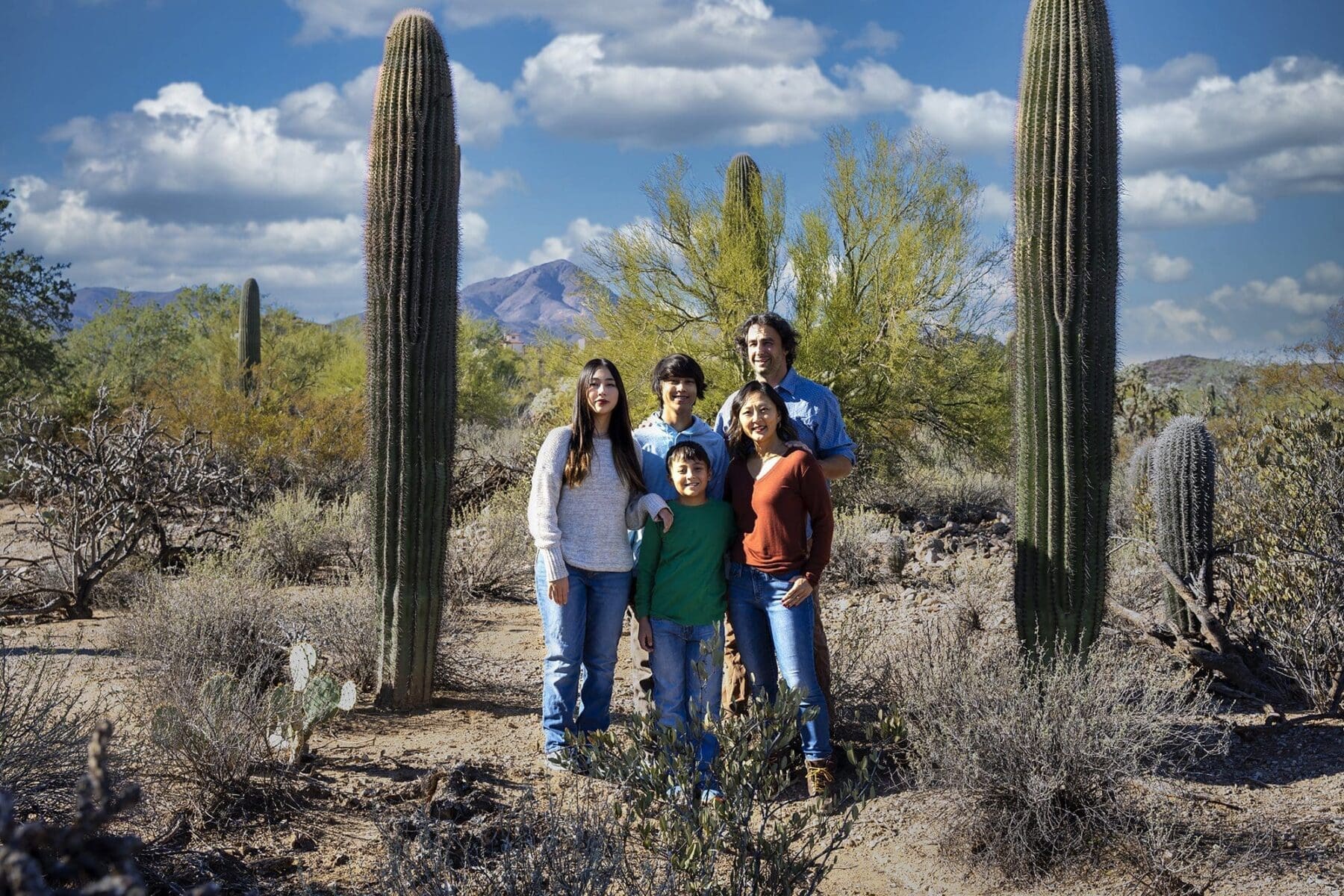 The Simpkins family stands together between two tall saguaros with a mountain and blue sky beyond.