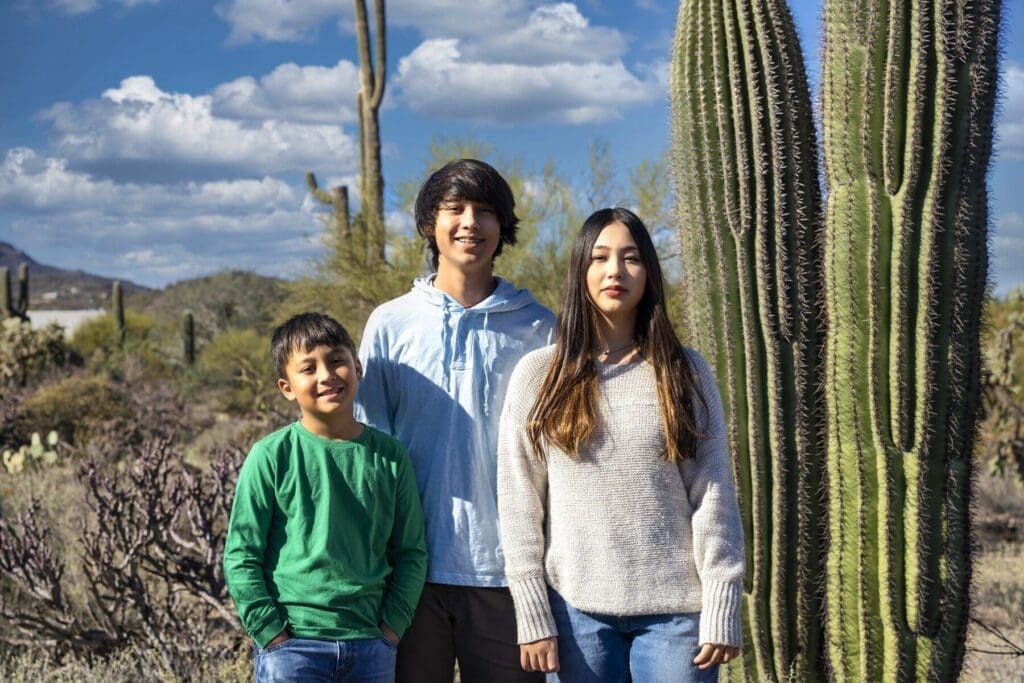 Three younger members of the Simpkins family stand together beside a tall saguaro under a blue sky.