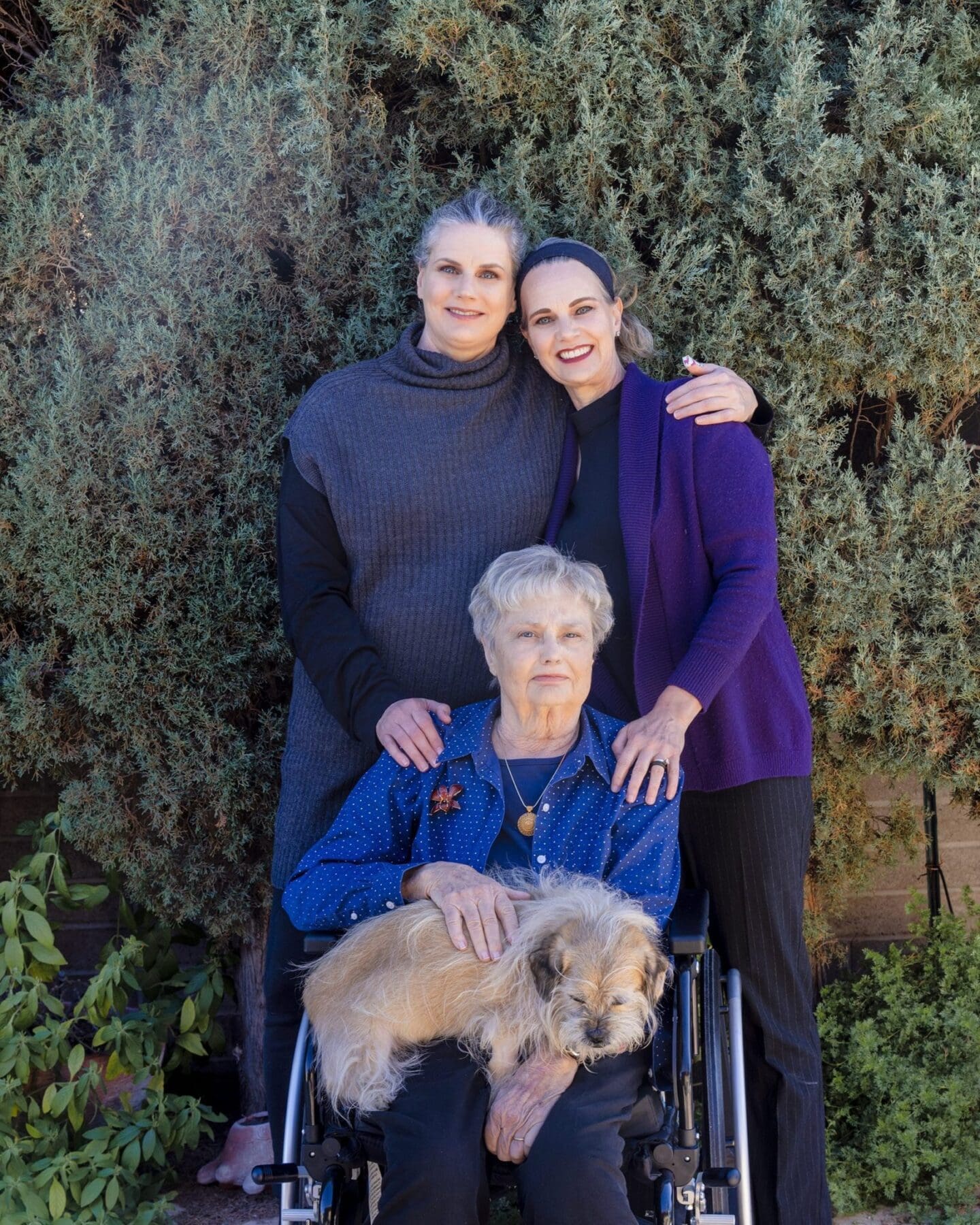 Three Slack family members pose together with a small dog in front of greenery.