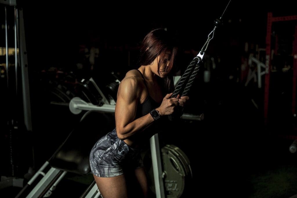 Clarissa leans forward while performing a rope pushdown in the gym.