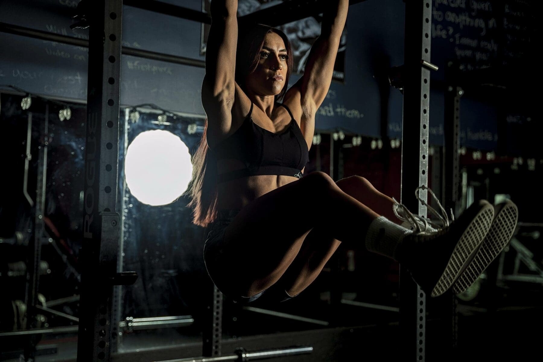 Clarissa raises her knees while hanging from a pull-up bar under dramatic light.