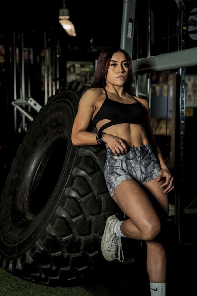 Clarissa leans against a large tractor tire in a vertical gym portrait.