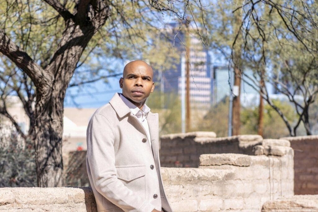 Aaron Owens faces the camera in a light suit with an adobe wall and desert trees behind him.