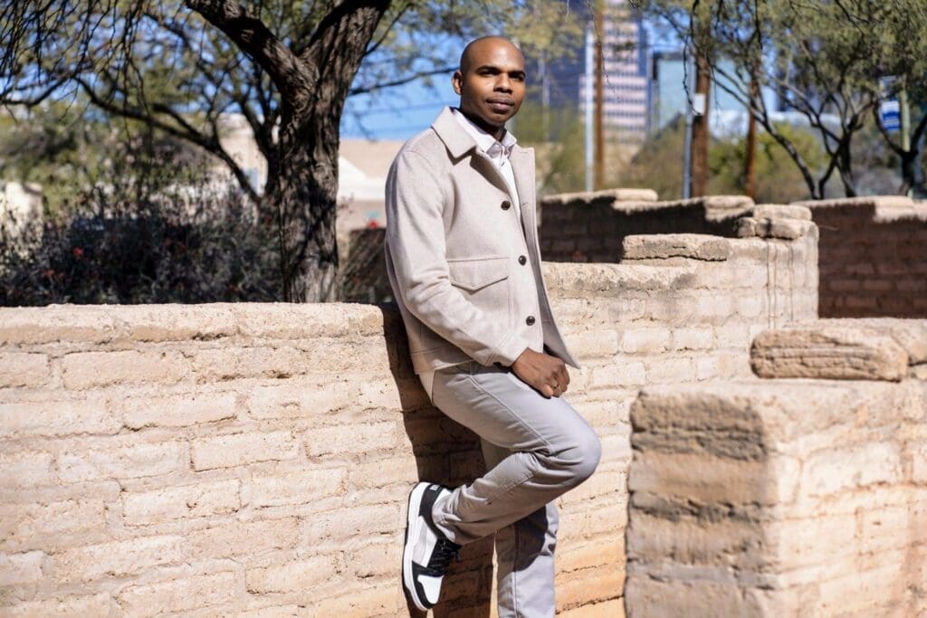 Aaron Owens leans against a low adobe wall in a light suit during an outdoor model session.