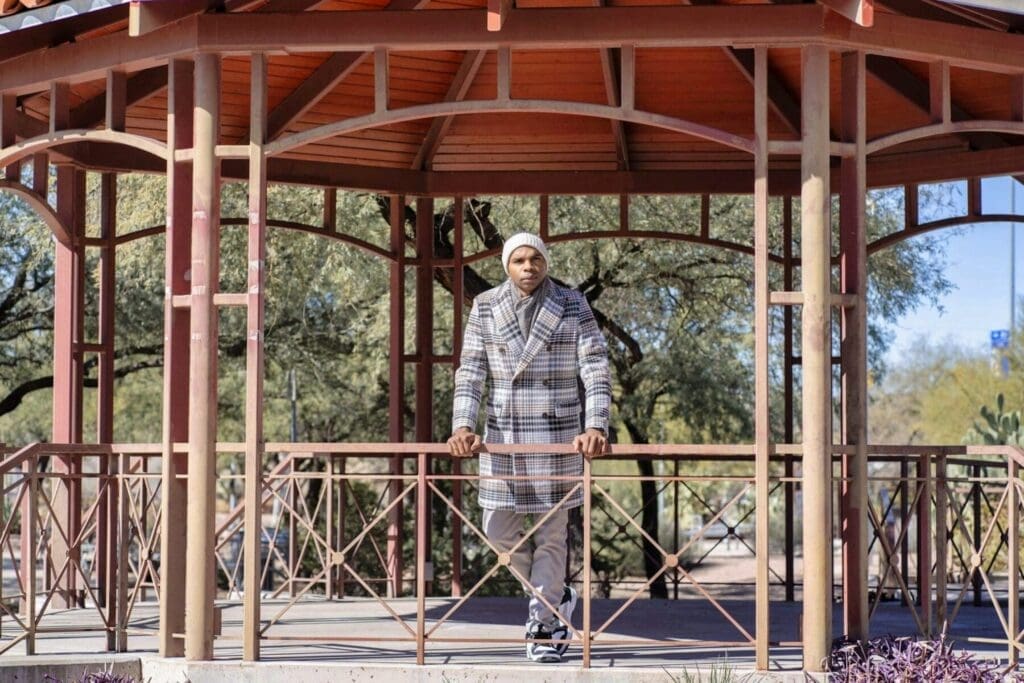 Aaron Owens stands symmetrically inside a red open-air pavilion wearing a plaid overcoat and knit cap.