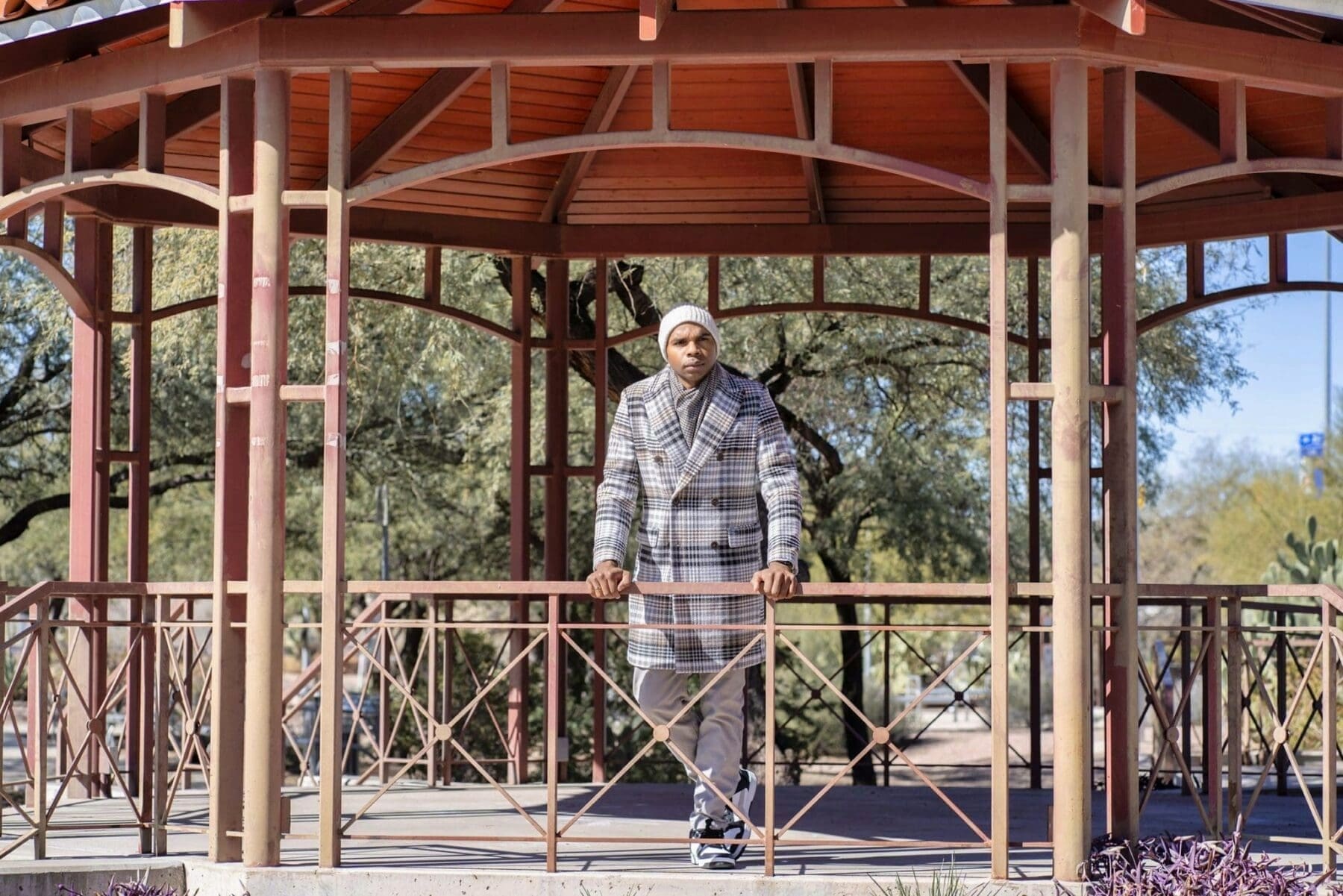 Aaron Owens stands symmetrically inside a red open-air pavilion wearing a plaid overcoat and knit cap.