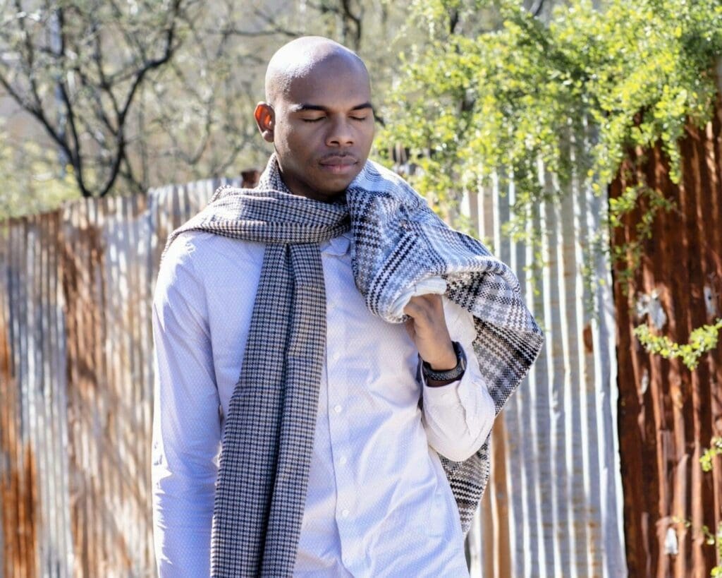 Aaron Owens looks down in a close portrait with a scarf over his shoulder and weathered metal behind him.