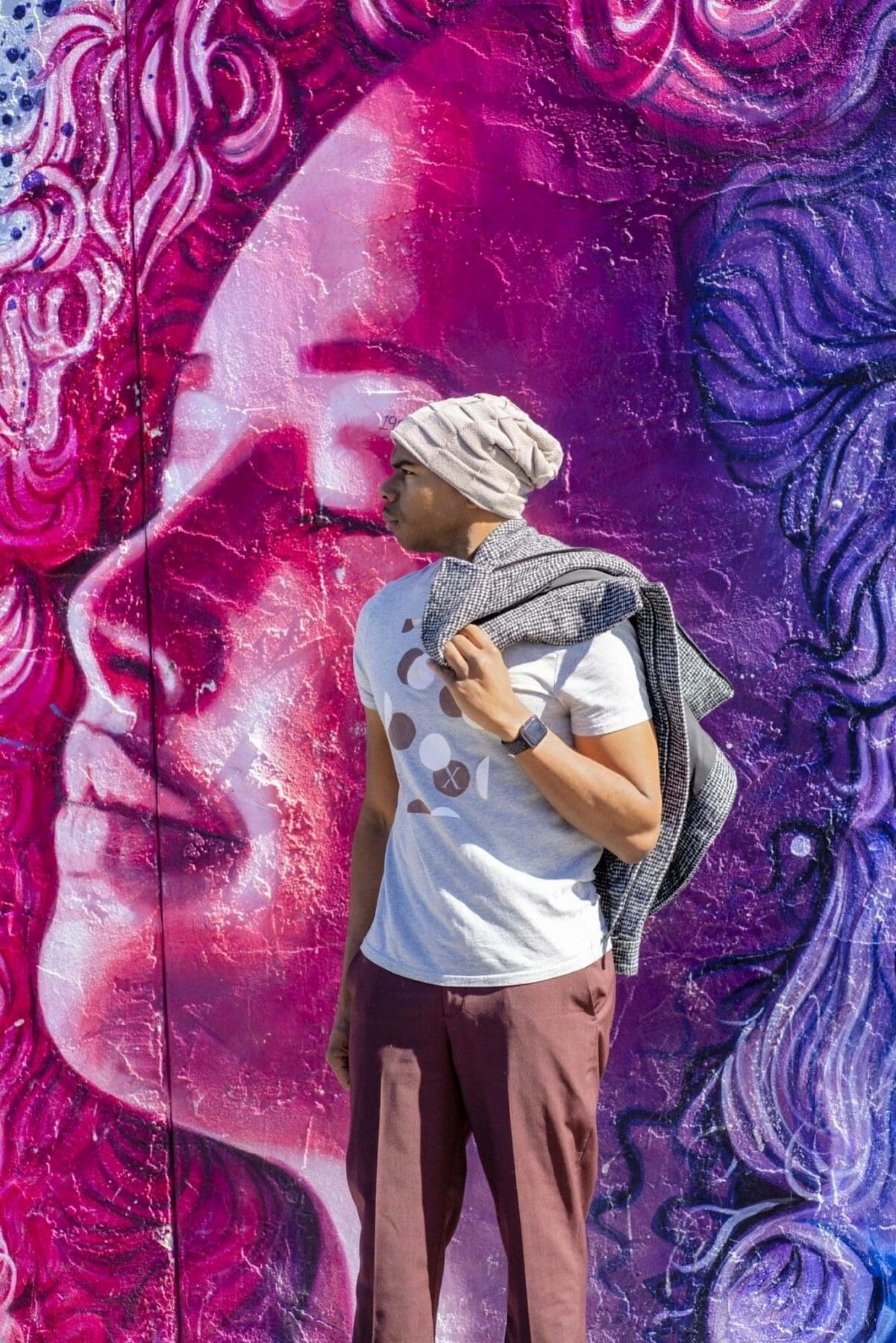 Aaron Owens stands in profile before a vivid mural of a face painted in magenta, purple, and blue.