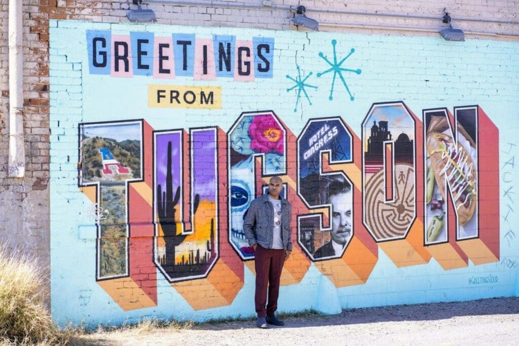 Aaron Owens stands in a gray jacket before a colorful wall mural reading Greetings from Tucson.
