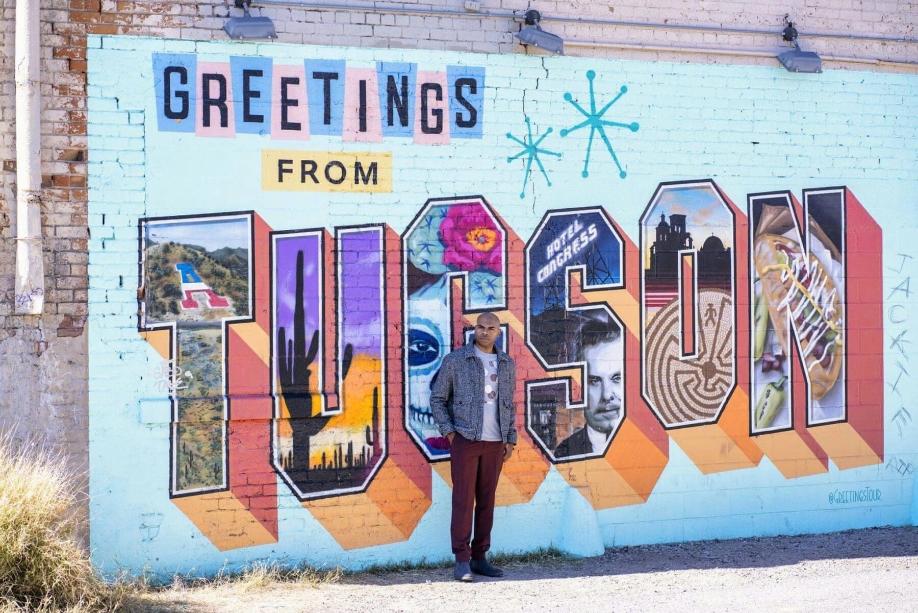 Aaron Owens stands in a gray jacket before a colorful wall mural reading Greetings from Tucson.