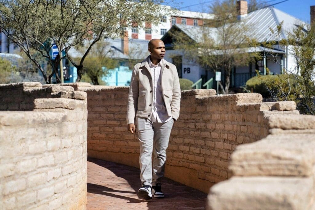 Aaron Owens walks toward the camera in a light suit between layered adobe walls.