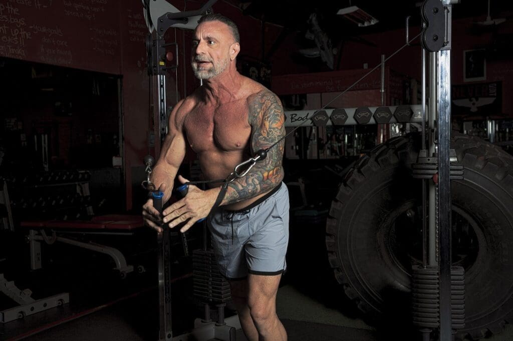 Demian Kolff performs a standing cable exercise in a dark gym, framed from the front with both handles visible.