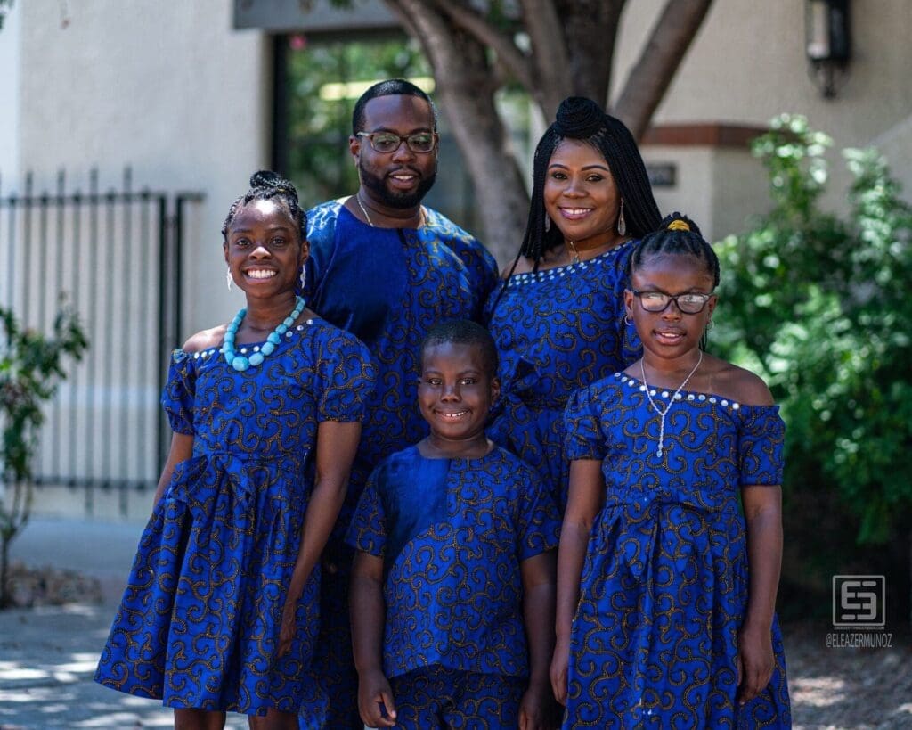 A large family group poses together beneath a courtyard tree in coordinated blue clothing.