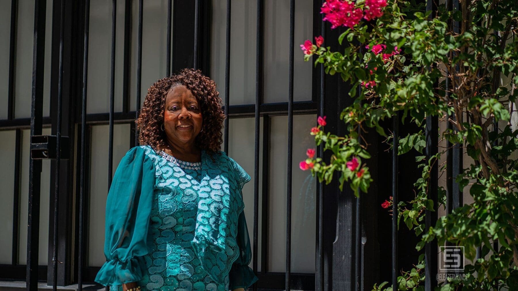 A woman in teal smiles beside a dark doorway and flowering bougainvillea.