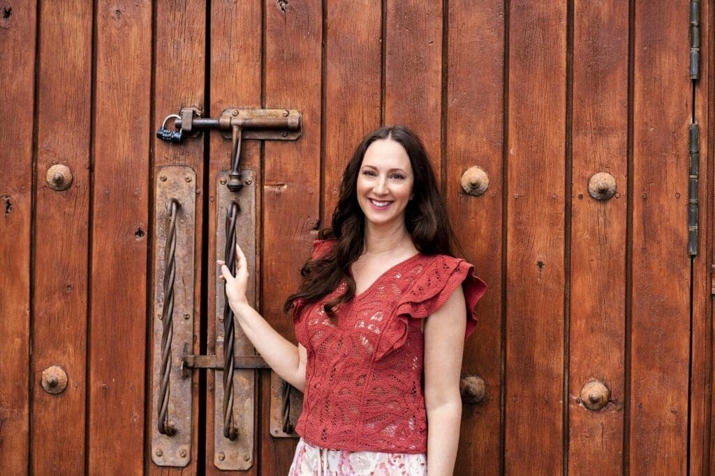 J smiles beside a large rustic wooden door while wearing a red ruffled blouse.