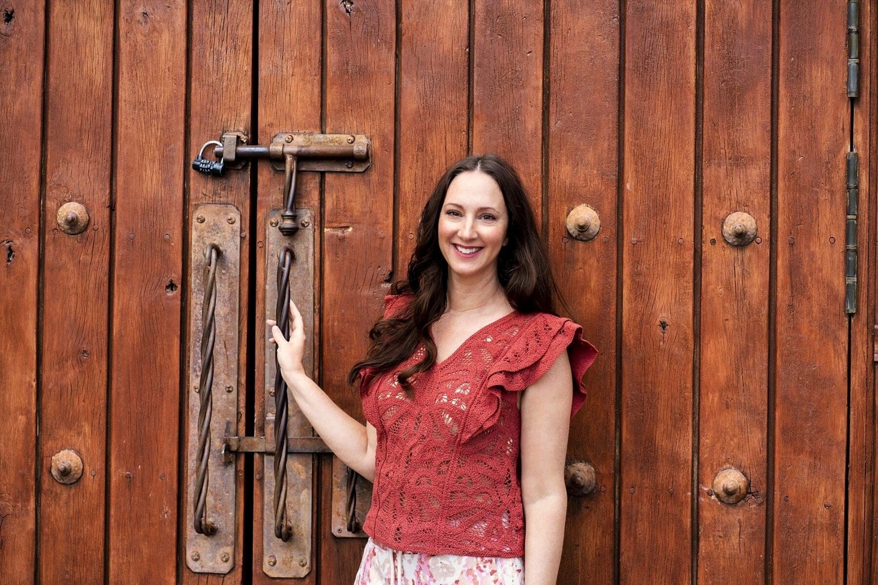 J smiles beside a large rustic wooden door while wearing a red ruffled blouse.