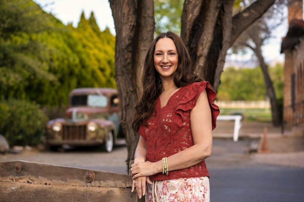 J leans on a rustic wooden fence with trees, lawn, and an older vehicle behind her.