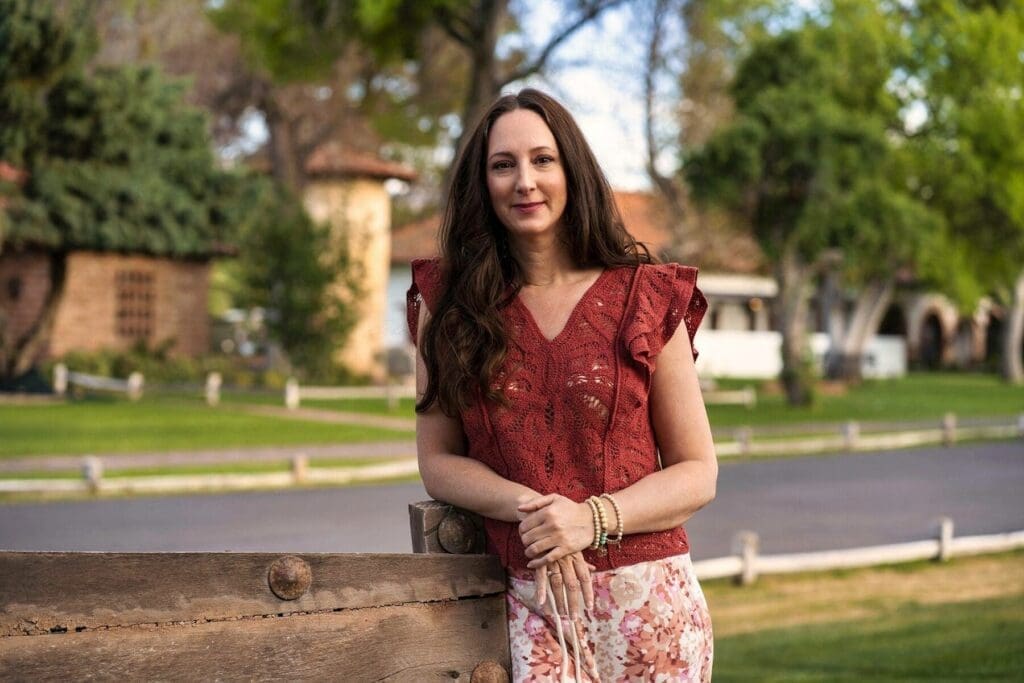 J faces the camera while resting her hands on a rustic wooden fence.