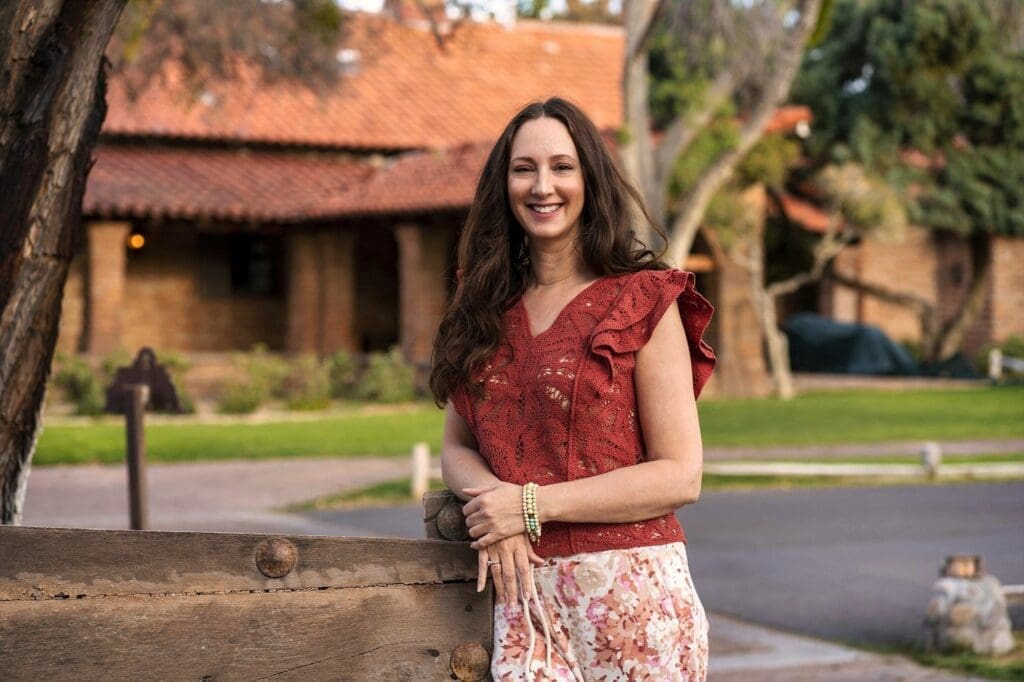 J stands beside a rustic fence with tiled-roof buildings and trees behind her.