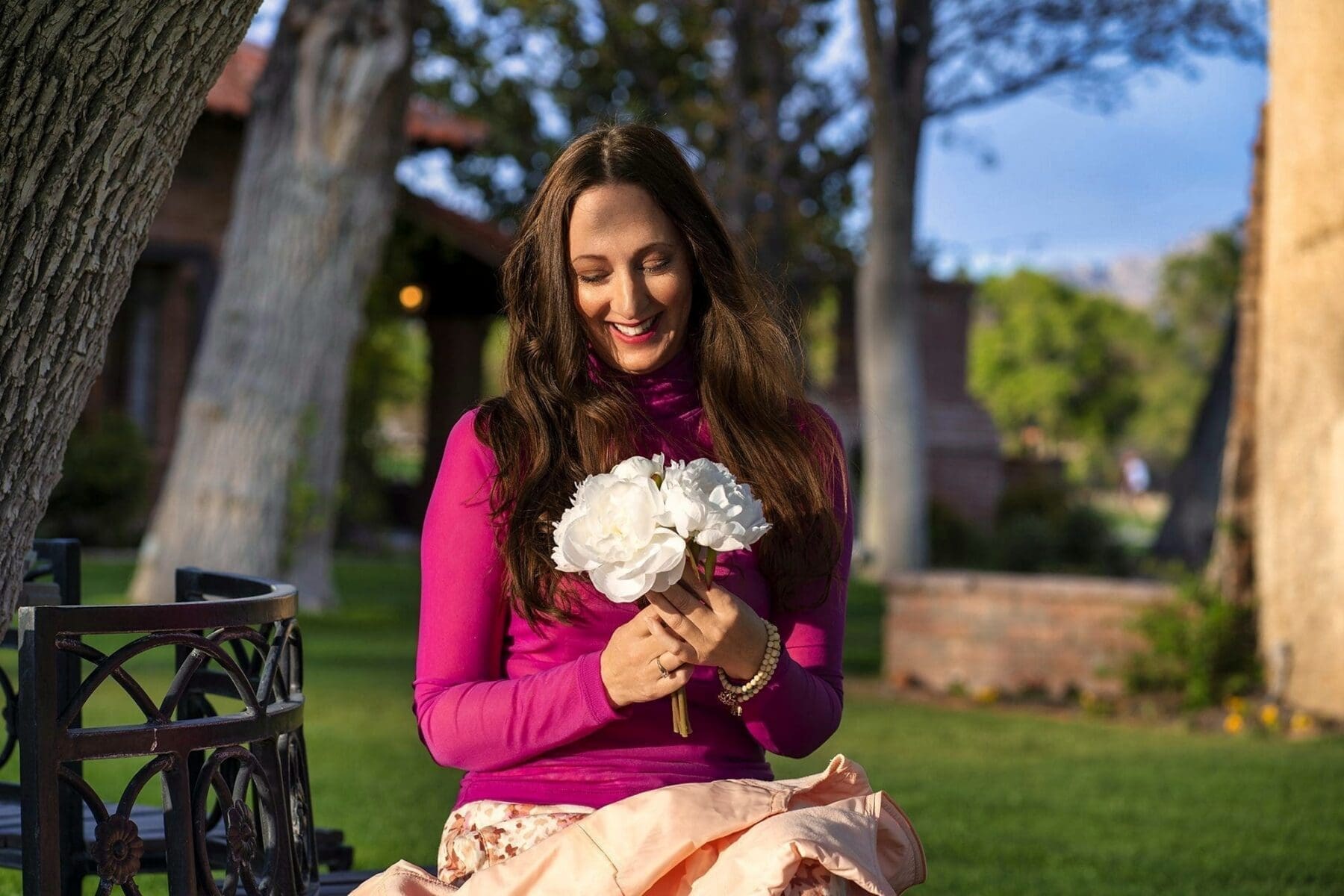 J looks down at a small bouquet of white flowers while seated outdoors in a magenta top.
