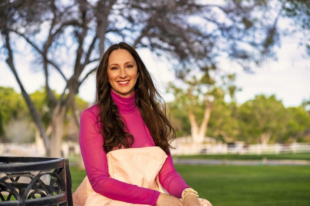 J smiles toward the camera while seated outdoors beneath spreading trees.