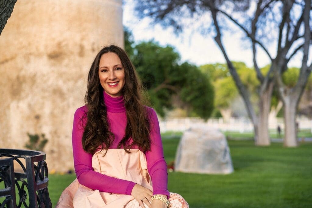 J sits near a sunlit stone wall with trees and lawn extending behind her.