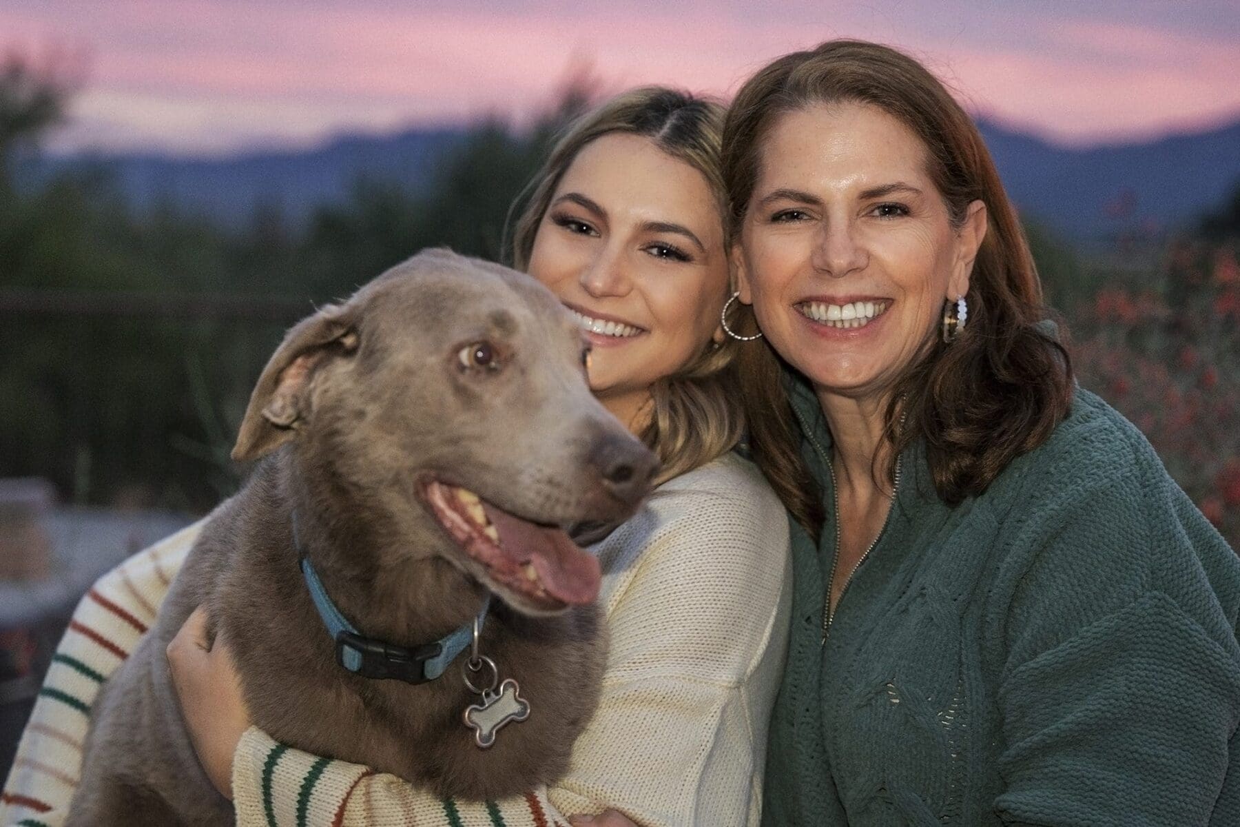 Two women smile close together while holding a gray dog, with a pink evening sky and mountains behind them.