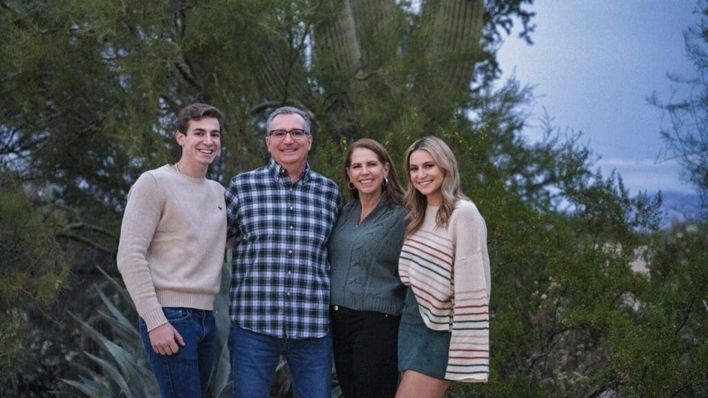 Four family members smile together in a close horizontal portrait framed by desert trees and a saguaro.