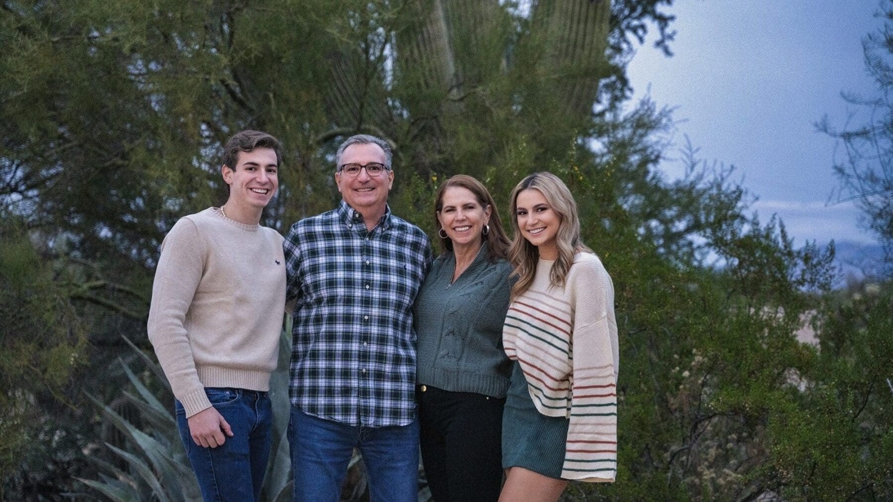 Four family members smile together in a close horizontal portrait framed by desert trees and a saguaro.