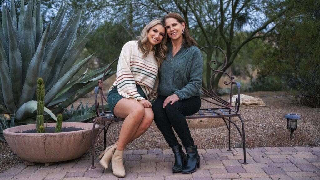 Two women sit closely together on a metal bench beside an agave and cactus planter.