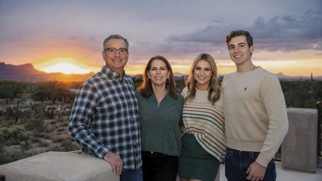 Four family members stand closely together on a terrace with the setting sun and mountains behind them.