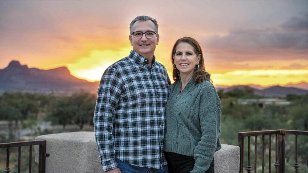 A man and woman stand together on a terrace with a vivid desert sunset and mountains behind them.