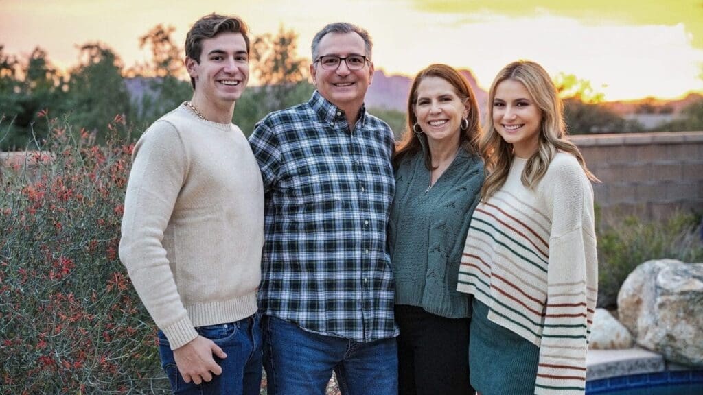 Four family members smile together outdoors beside flowering shrubs, rocks, and the edge of a pool.