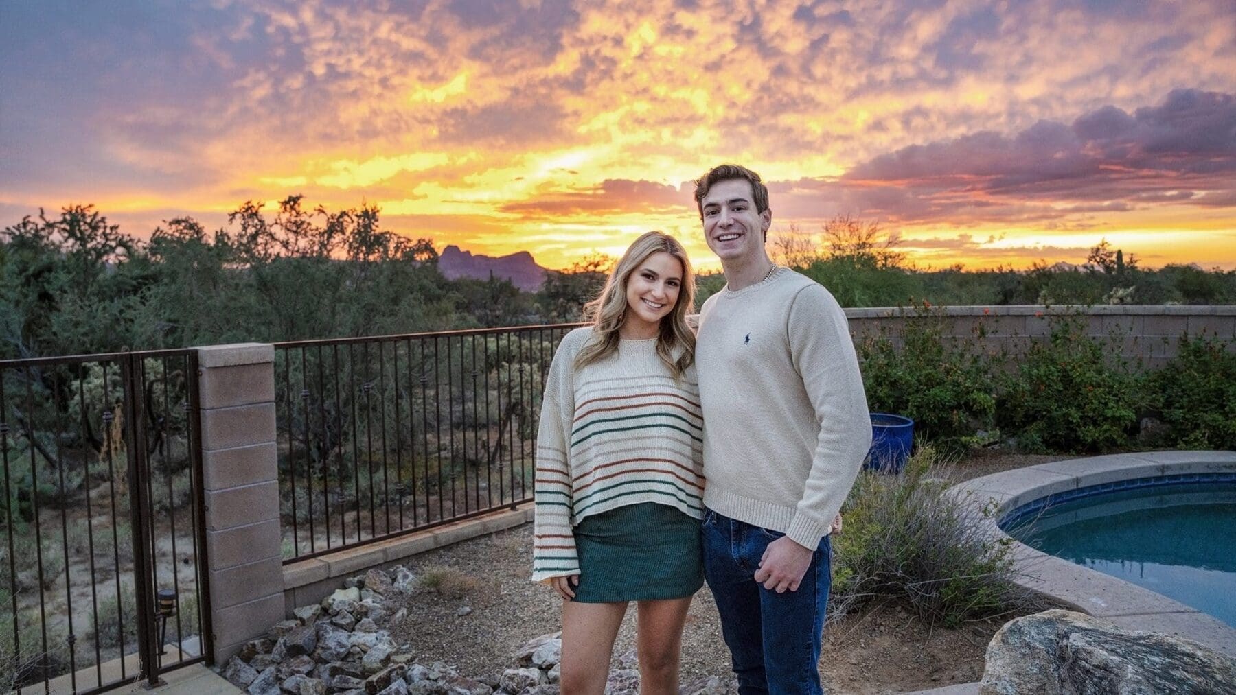 A young woman and young man stand together near a pool beneath a vivid orange and purple evening sky.