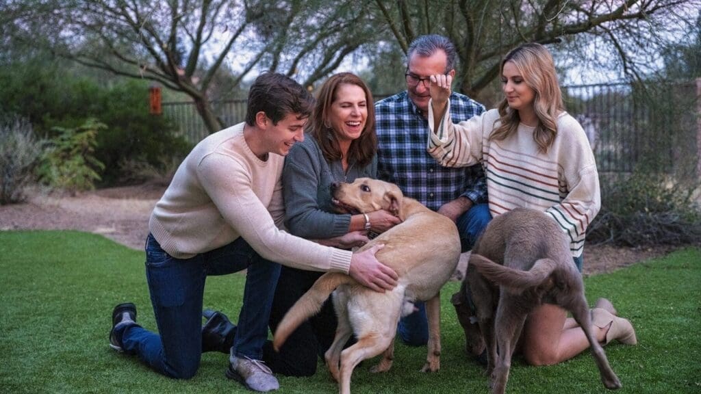 Four family members laugh and interact with two dogs while kneeling together on a lawn.