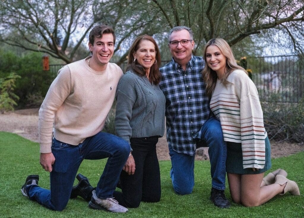 Four family members kneel closely together on a lawn with desert trees behind them.