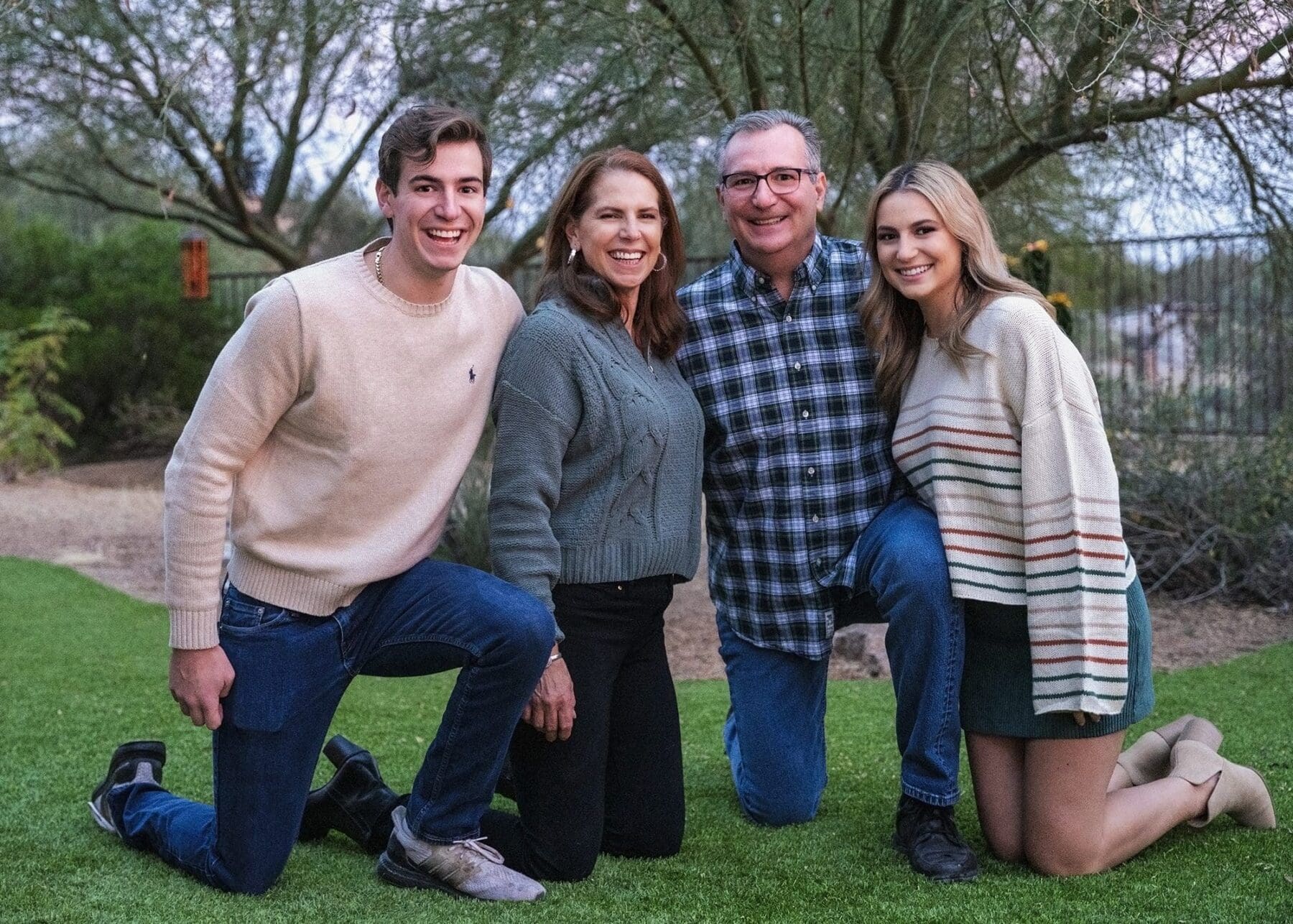 Four family members kneel closely together on a lawn with desert trees behind them.
