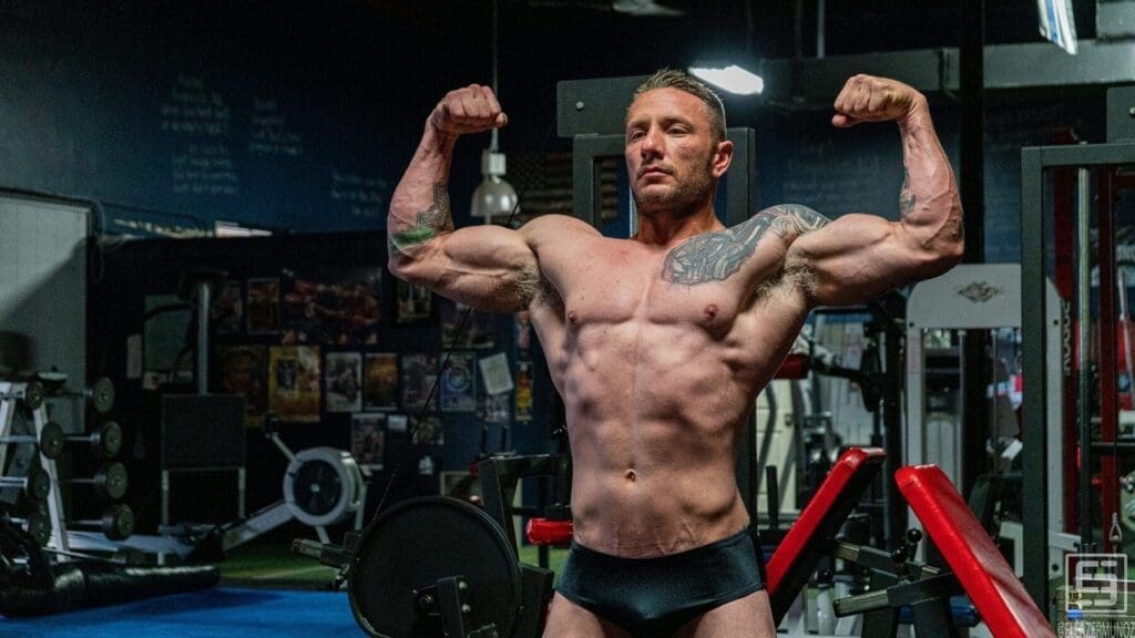Irish Michael performs a front double-biceps pose in black posing trunks inside the gym.