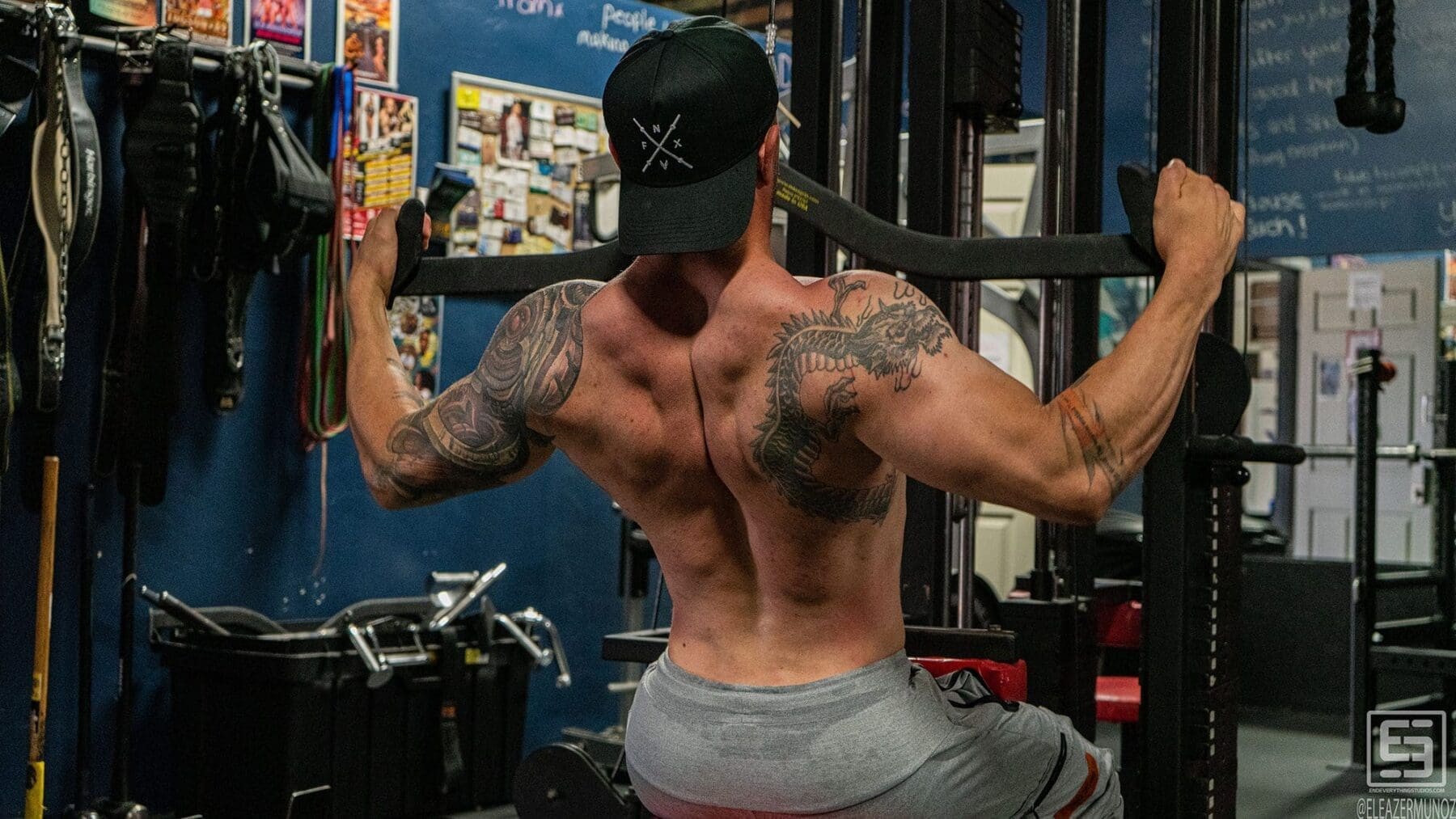 Irish Michael performs a seated lat pulldown viewed from behind, showing his back tattoos and muscle definition.