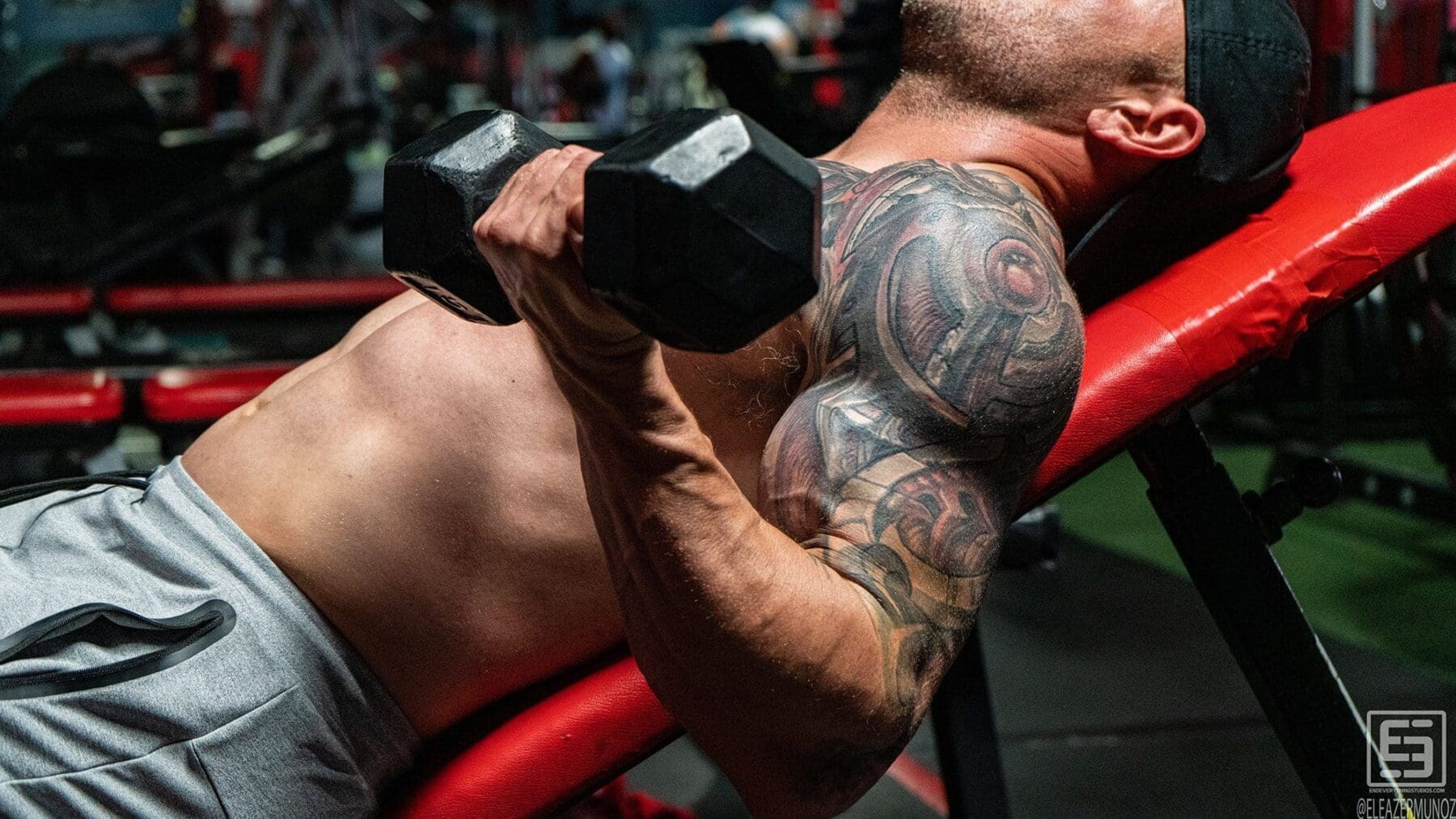 Irish Michael performs an incline dumbbell curl on a red bench in a tight upper-body frame.